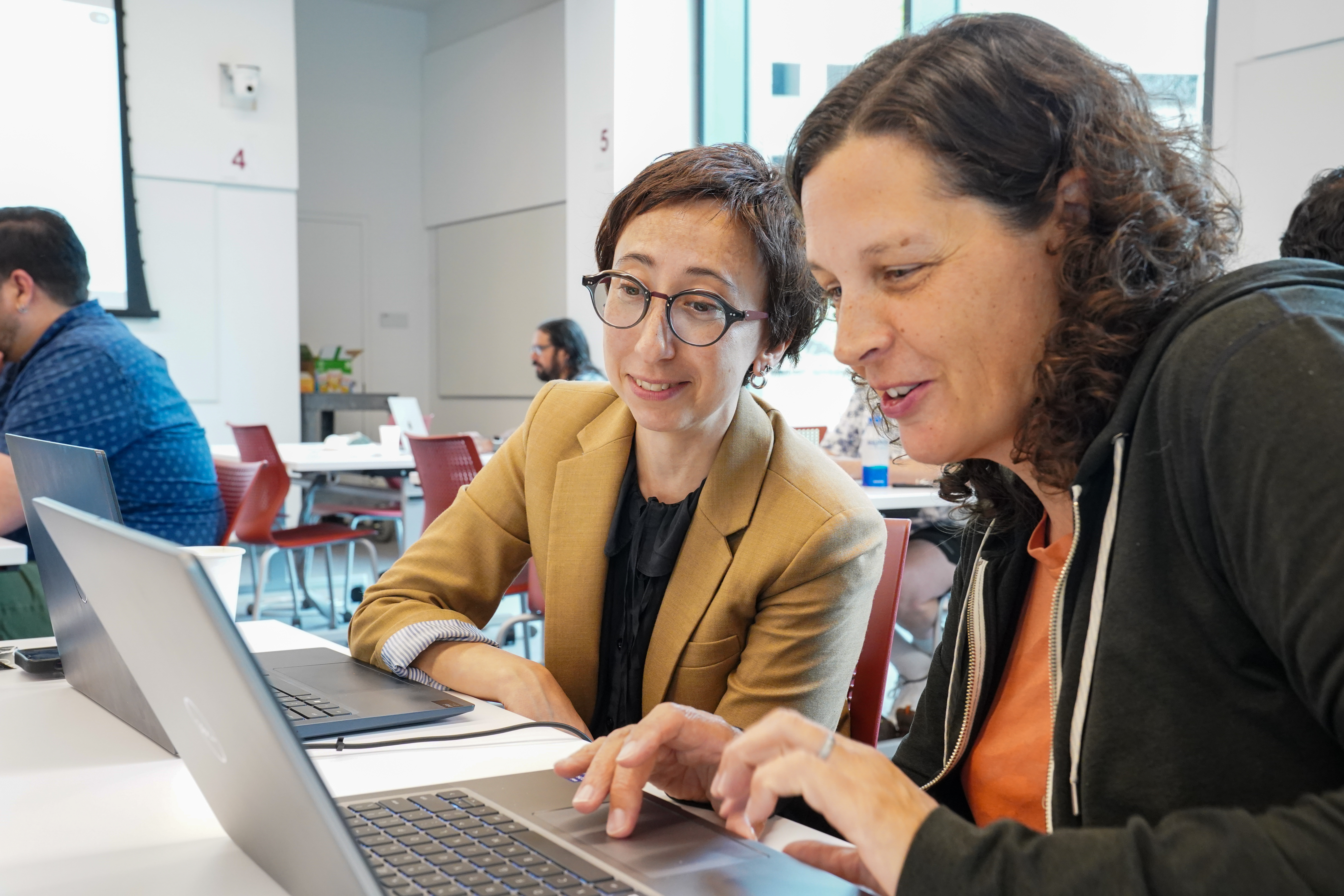 two women with brown hair type on two laptops and work 