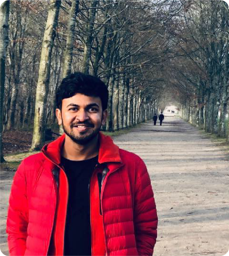 A color photo of a man wearing a red jacket along a tree-lined road.