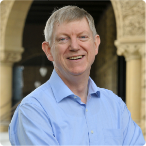 A color photo of a man smiling for a photo outside wearing a blue dress shirt.