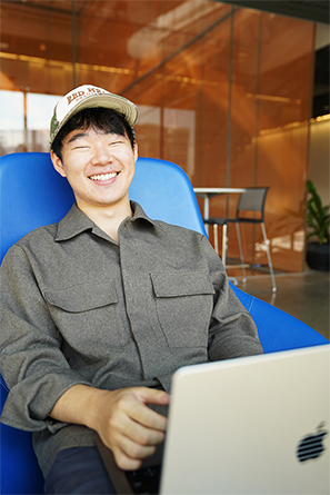 A color photo of a student sitting in a blue chair wearing a baseball cap.