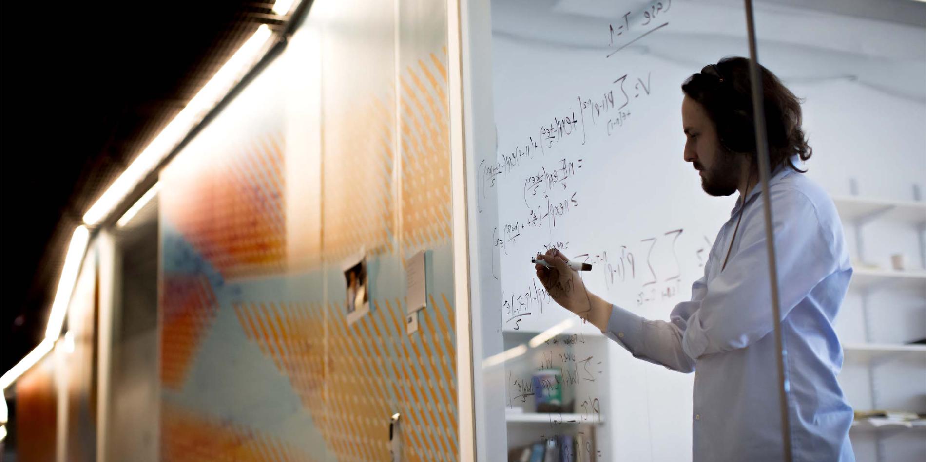 Person writing equations on a glass wall in a research setting.