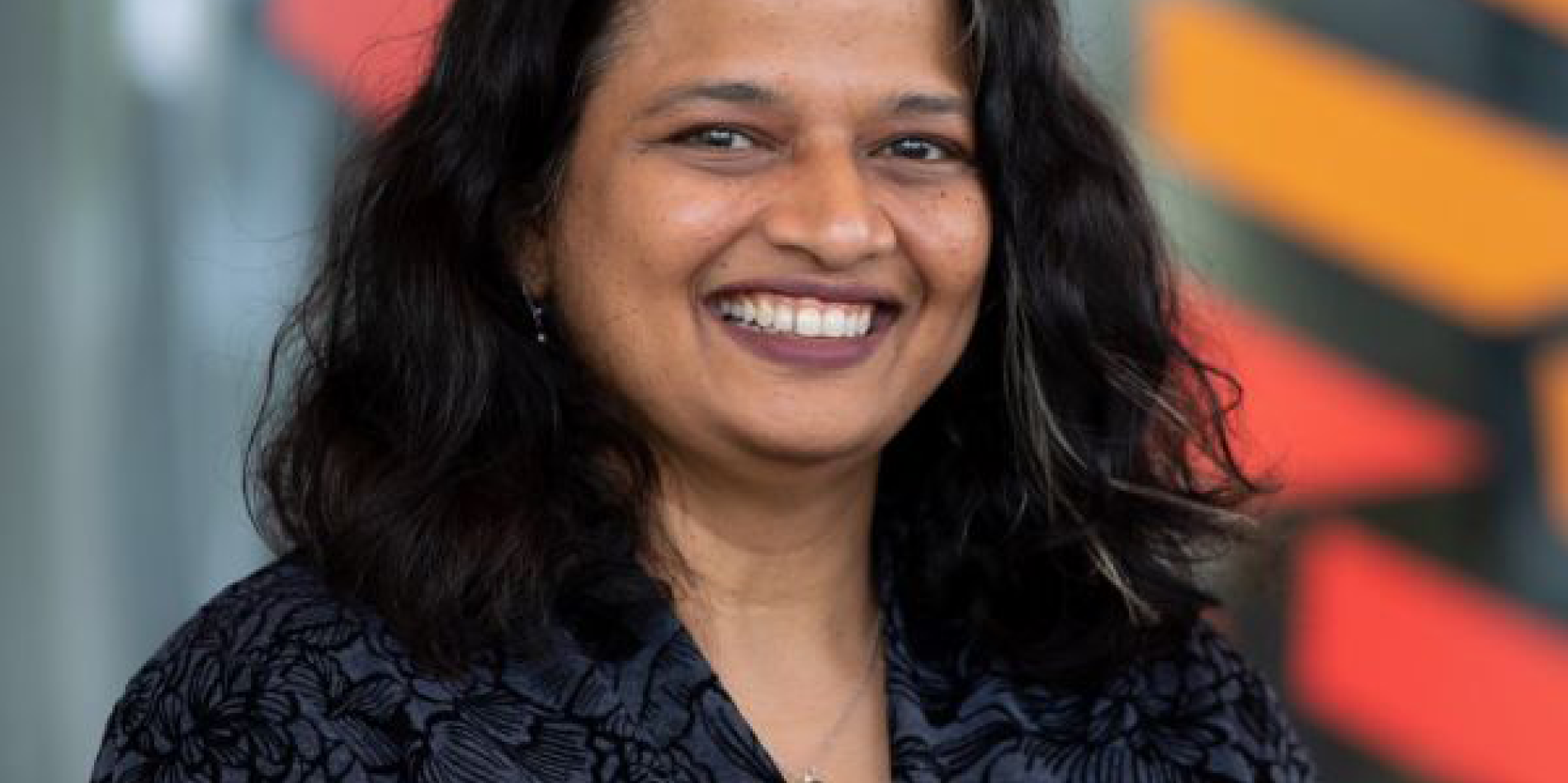 dark haired woman with dark hair smiles in front of a building with red and orange streaks