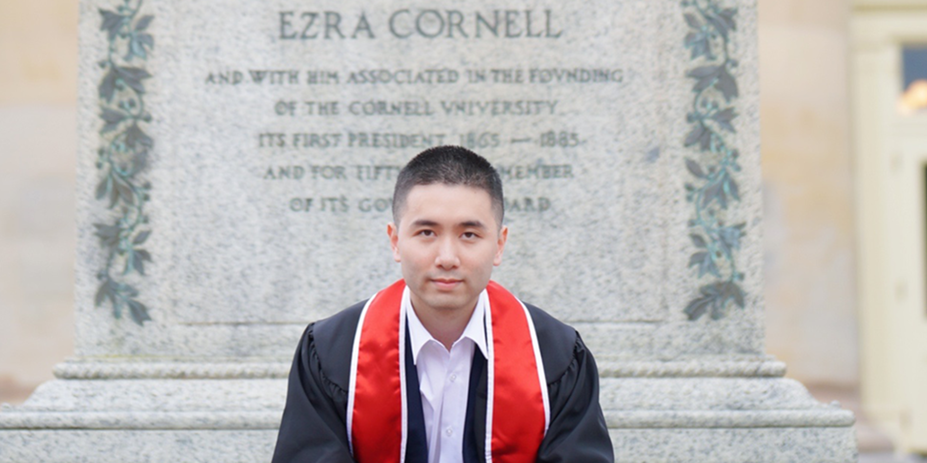 a young asain man sits on a gray statue wearing a black graduation robe and red 