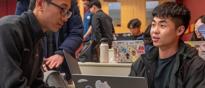 Two students talk across a laptop