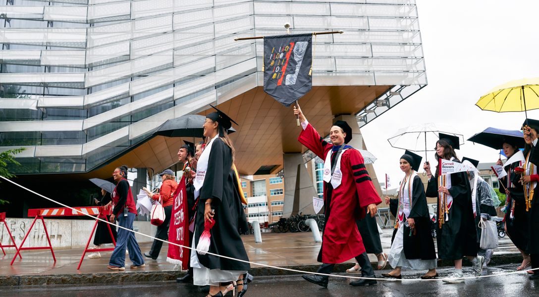 A color photo showing people walking to Cornell University graduation behind the Cornell Bowers flag