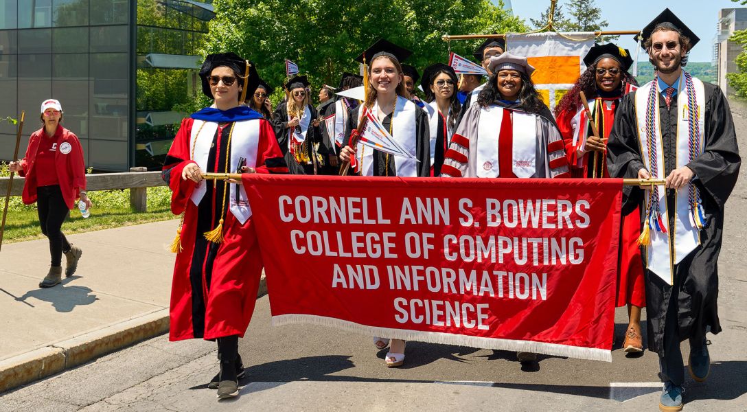 Several students in graduation robes march down a street on sunny day holding a banner that says Cornell Ann S. Bowers College of Computing and Information Science.