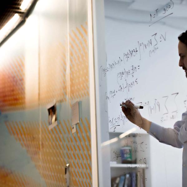 Person writing equations on a glass wall in a research setting.