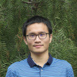 A color photo of a man smiling for a photo in front of green trees.