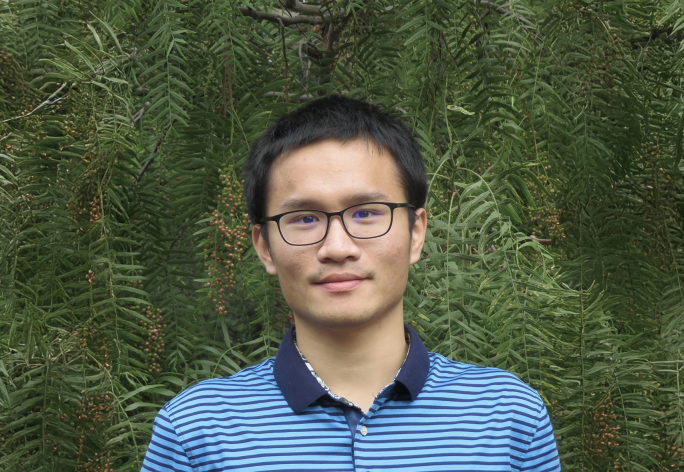 A color photo of a man smiling for a photo in front of green trees.