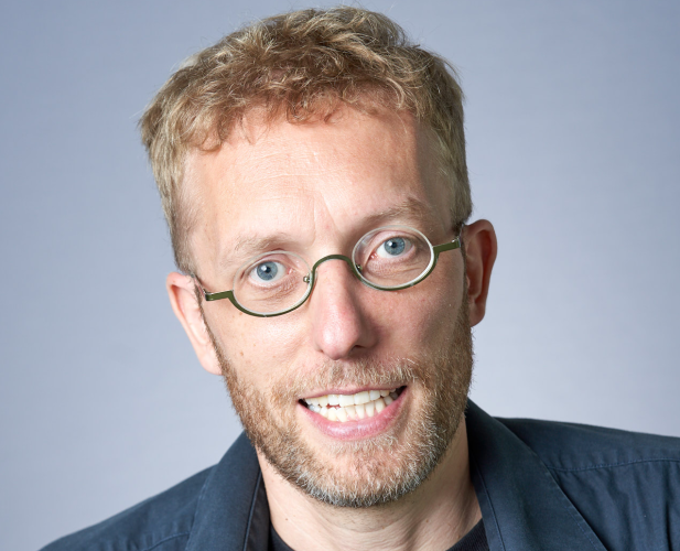 A photo of Kilian Weinberger, a man with curly blond hair, glasses, a beard and mustache, in a gray shirt in front of a gray background