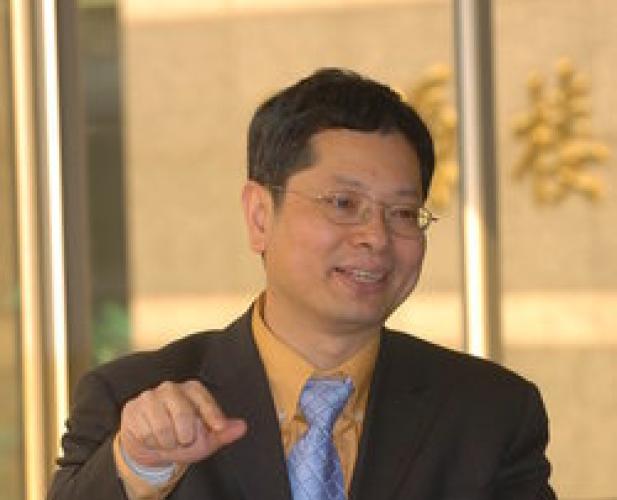 man talking, wearing a dark suit with blue tie and yellow shirt