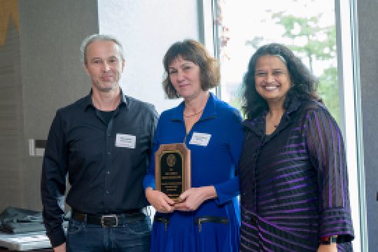 A color photo showing a man and two women posing for a photo. The woman in the middle is holding an award