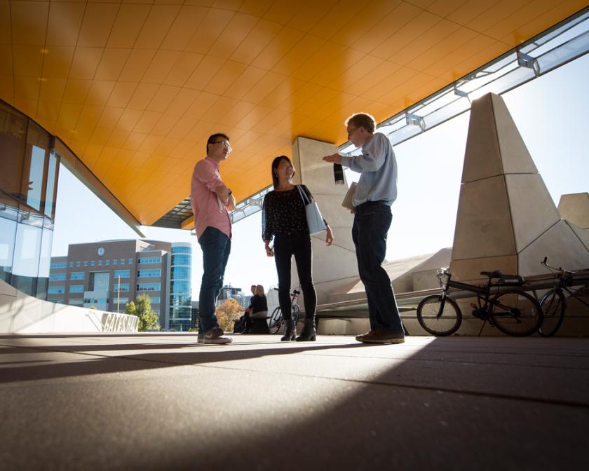 Three people standing under the orange overhang of Gates Hall on a sunny day