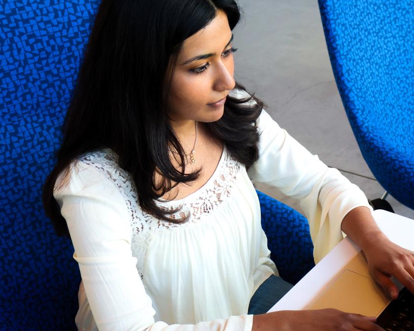 a young female student with tan skin and long black hair sits on a blue chair while typing on a laptop
