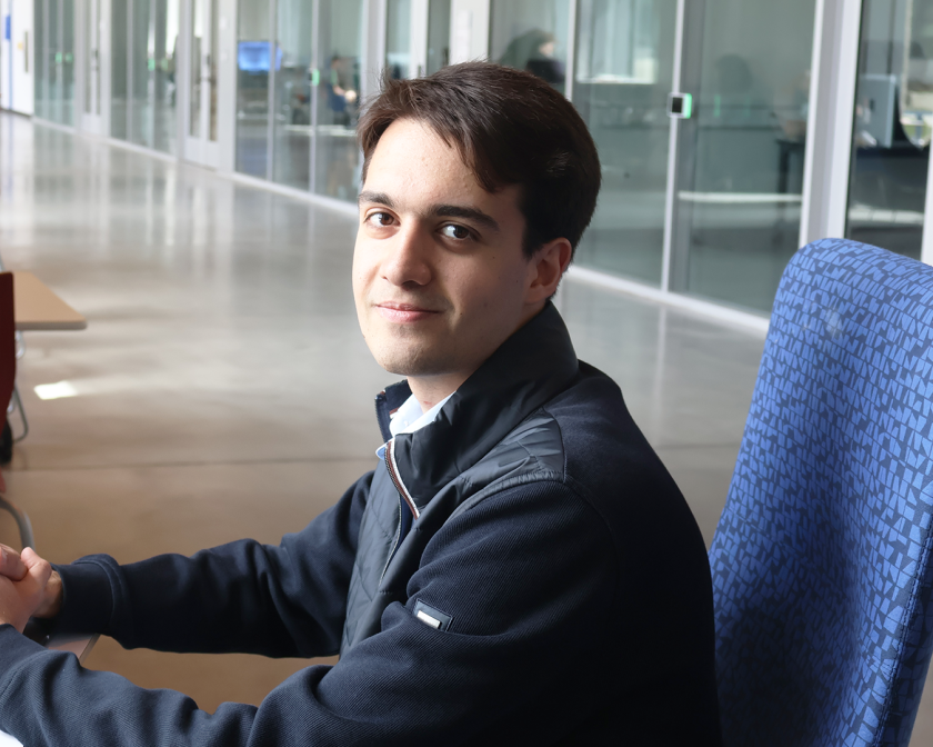 a young male student with short brown hair sits at a chair and works on a laptop