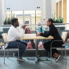 A color photo of two people sitting at a table, speaking with each other