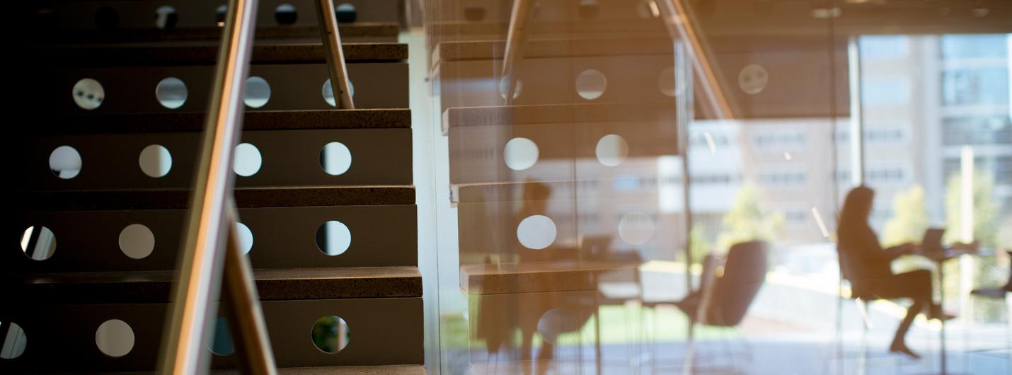 a metal staircase on the left forms into a glass wall reflecting building angles and orange light with two people working at tables in the background