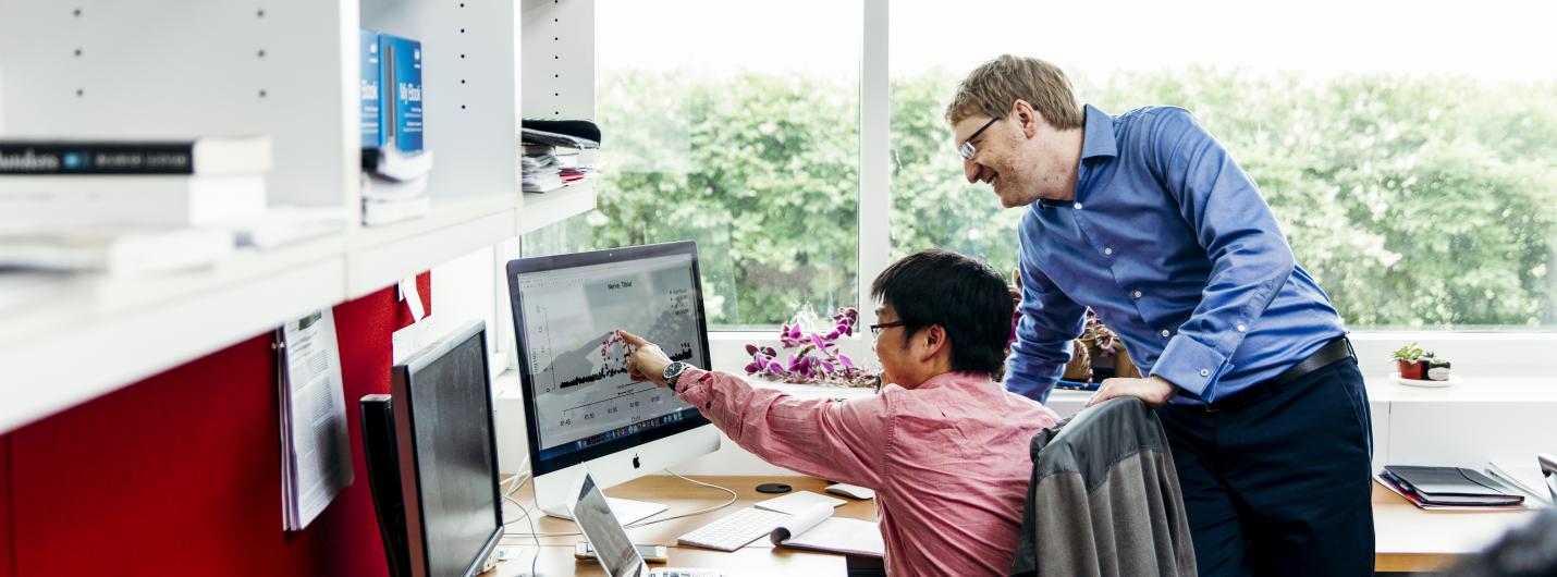 a student sits at a desk pointing a desktop while a male faculty member looks at the screen behind him